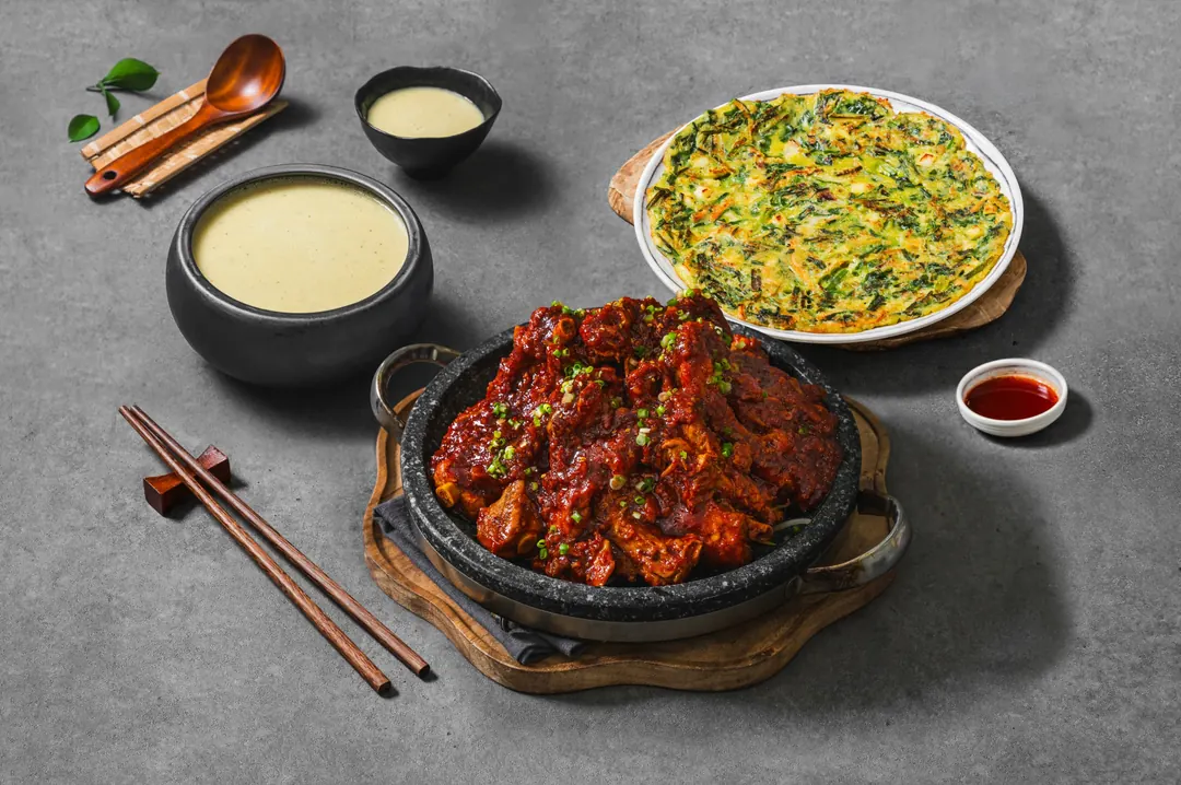 돌판에 담긴 양념 등갈비구이와 부쭈전, 뽀얀 막걸리, 수저와 소스가 회색 테이블에 차려진 고봉당 한상차림